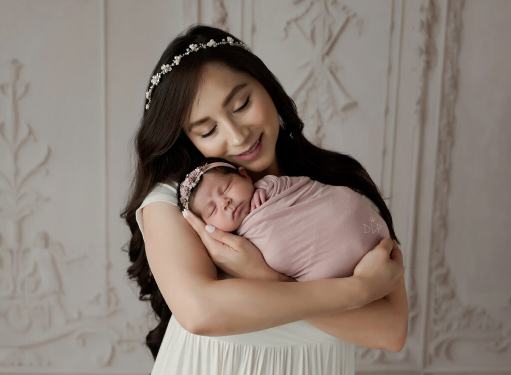Special offer by Austin Newborn Photographer Dazzling Light Photography. Mom in white dress holding baby girl that is wrapped in pink swaddle